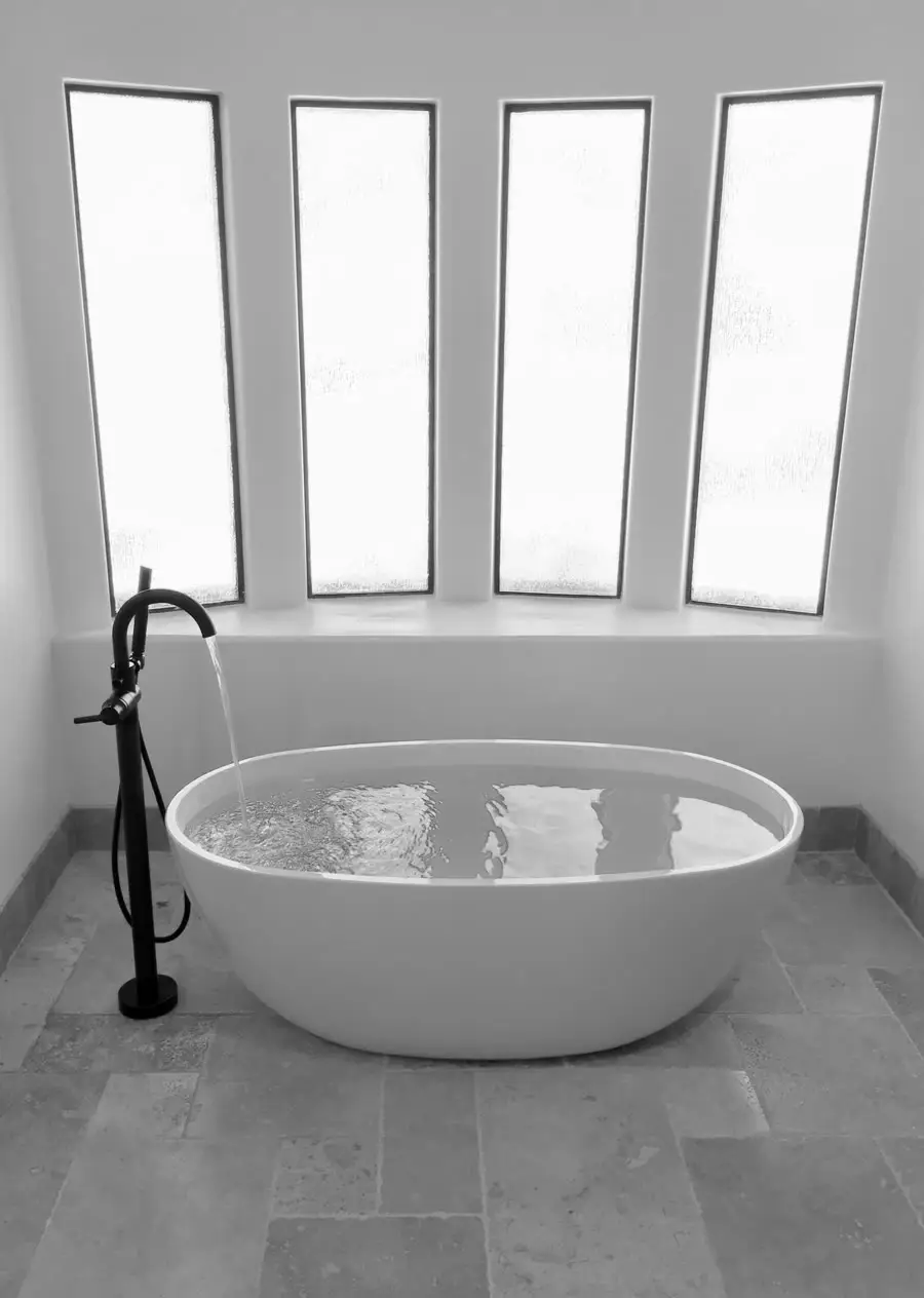 Freestanding soaking tub set under a run of tall narrow windows on a large-format stone tile floor